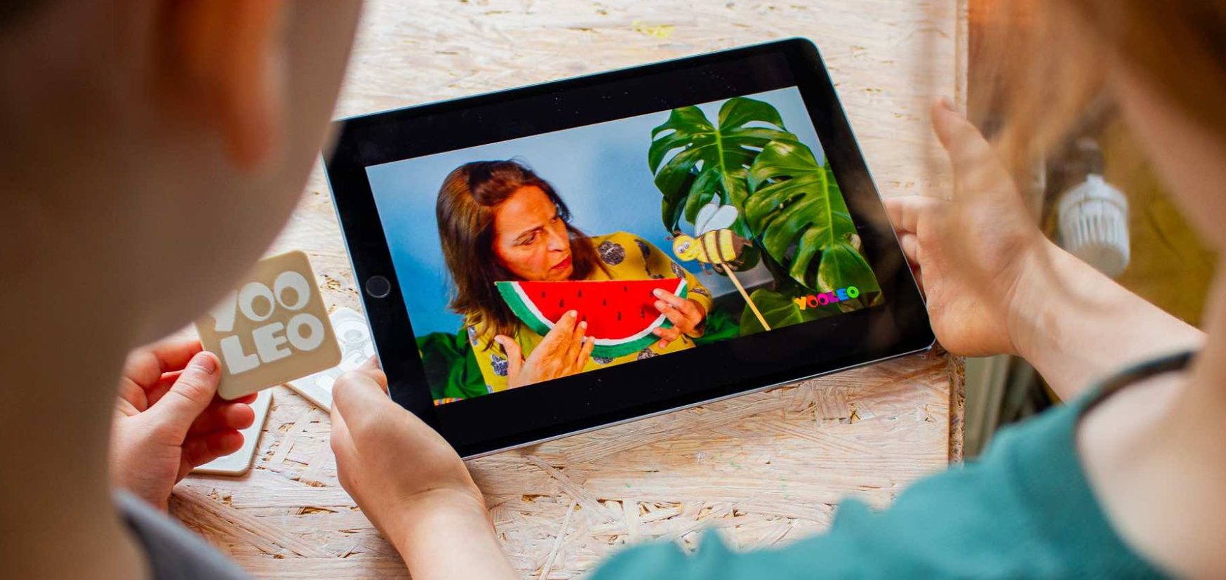 YOOLEO Lernplattform Zwei Kinder schauen gemeinsam ein Video auf einem Tablet. Im Video ist eine Frau mit Melone und Pflanzen zu sehen.